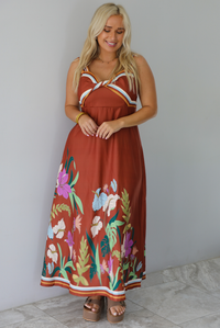 A woman wearing a rust colored maxi dress with other colors at the bust in a striped pattern, with a multi-colored floral pattern at the hem, standing indoors against a white wall with white marble flooring. 