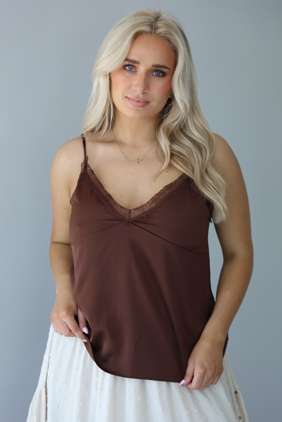 Woman wearing a satin brown tank that is untucked and paired with a high waisted sand colored midi length skirt. The photo is taken in front of a white wall.