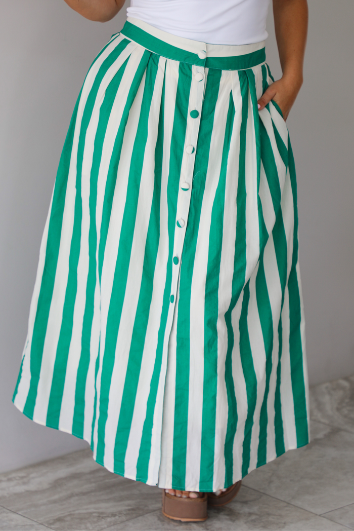 Closeup of a woman wearing a white tank top and a maxi length cream and green striped skirt, focused in on the skirt, which has a high waistline, functional pockets on either side, functional button closures in the front, and has a flowy fit, standing indoors against a white wall with white marble flooring. 
