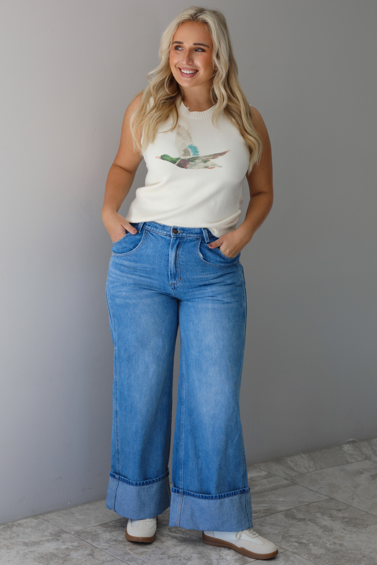 A woman wearing a cream colored sweater vest with a mallard duck on the front, paired with medium wash denim with wide leg hem, standing indoors against a white wall with white marble flooring. 