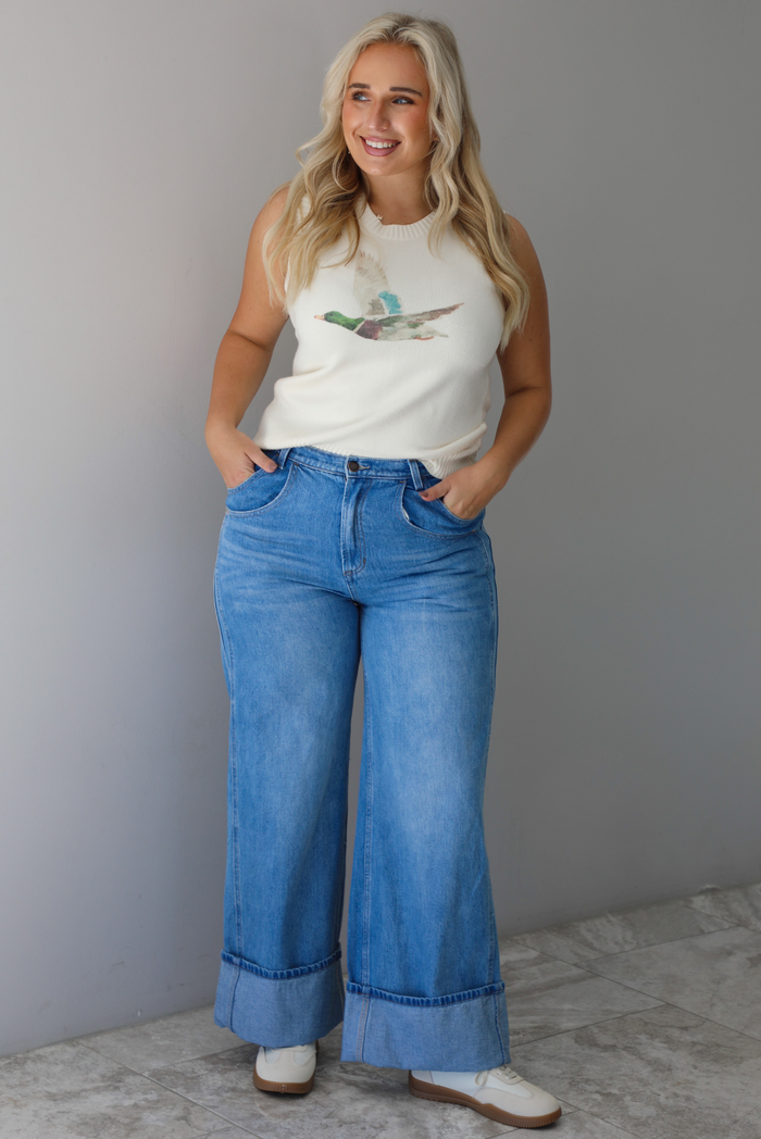 A woman wearing a cream colored sweater vest with a mallard duck on the front, paired with medium wash denim with wide leg hem, standing indoors against a white wall with white marble flooring. 