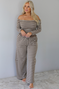 A woman wearing a brown with cream striped matching two piece set consisting of a long sleeved, off the shoulder top and high waisted pants, standing indoors against a white wall with white marble flooring. 