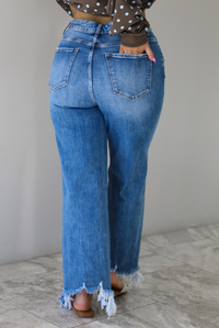 Woman wearing a medium wash denim that are high waisted, have functional front and back pockets, functional button and zipper closures, and have a raw, distressed hem. The photo shows the back of jeans, and the models hand is in the back right pocket. The photo is taken indoors against a white wall with white marble flooring.