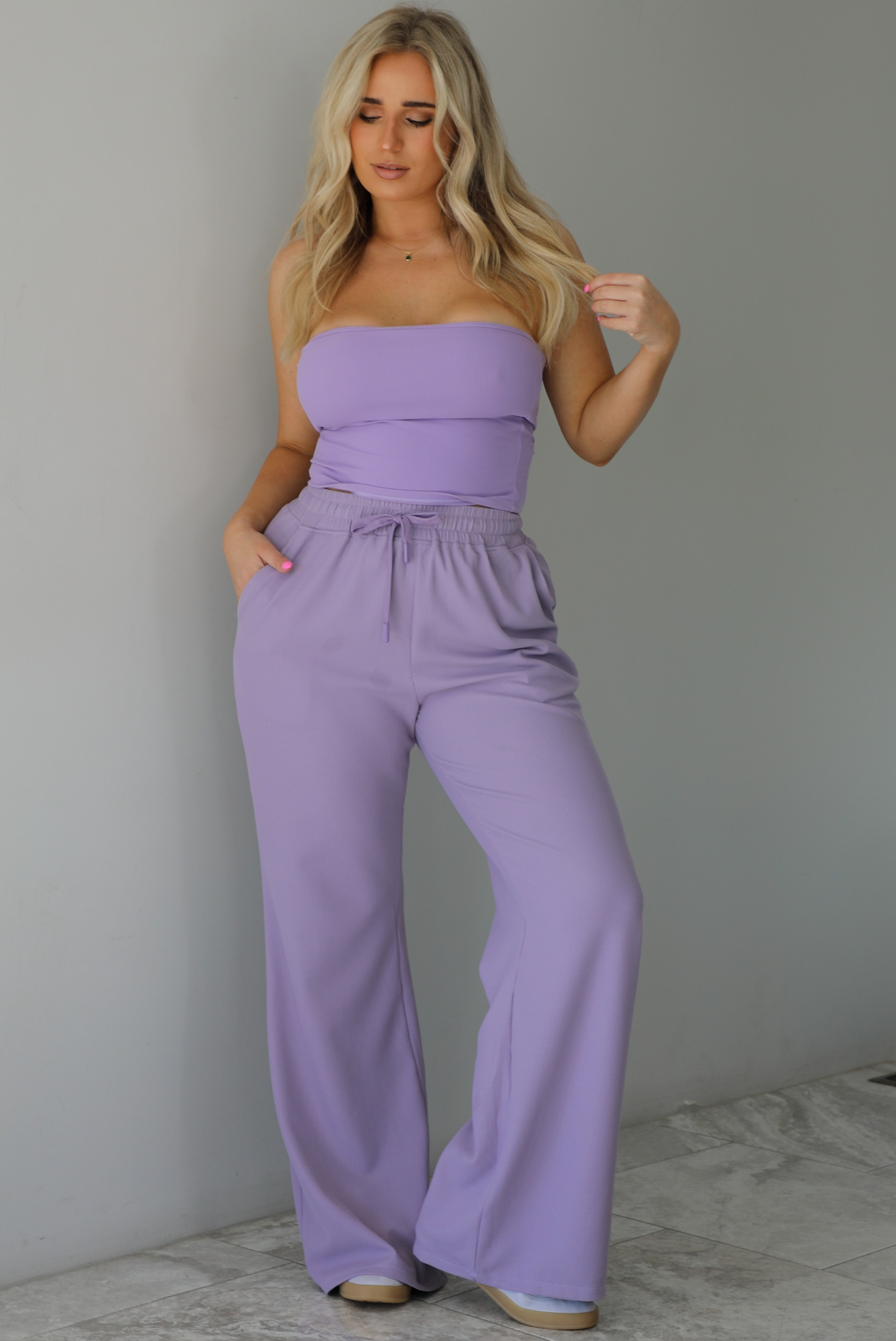 Woman wearing a two piece lavender set featuring a strapless, fitted tube top, and high waisted, flared hem pants. The photo is taken indoors against a white wall with white marble flooring.