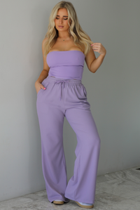 Woman wearing a two piece lavender set featuring a strapless, fitted tube top, and high waisted, flared hem pants. The photo is taken indoors against a white wall with white marble flooring.