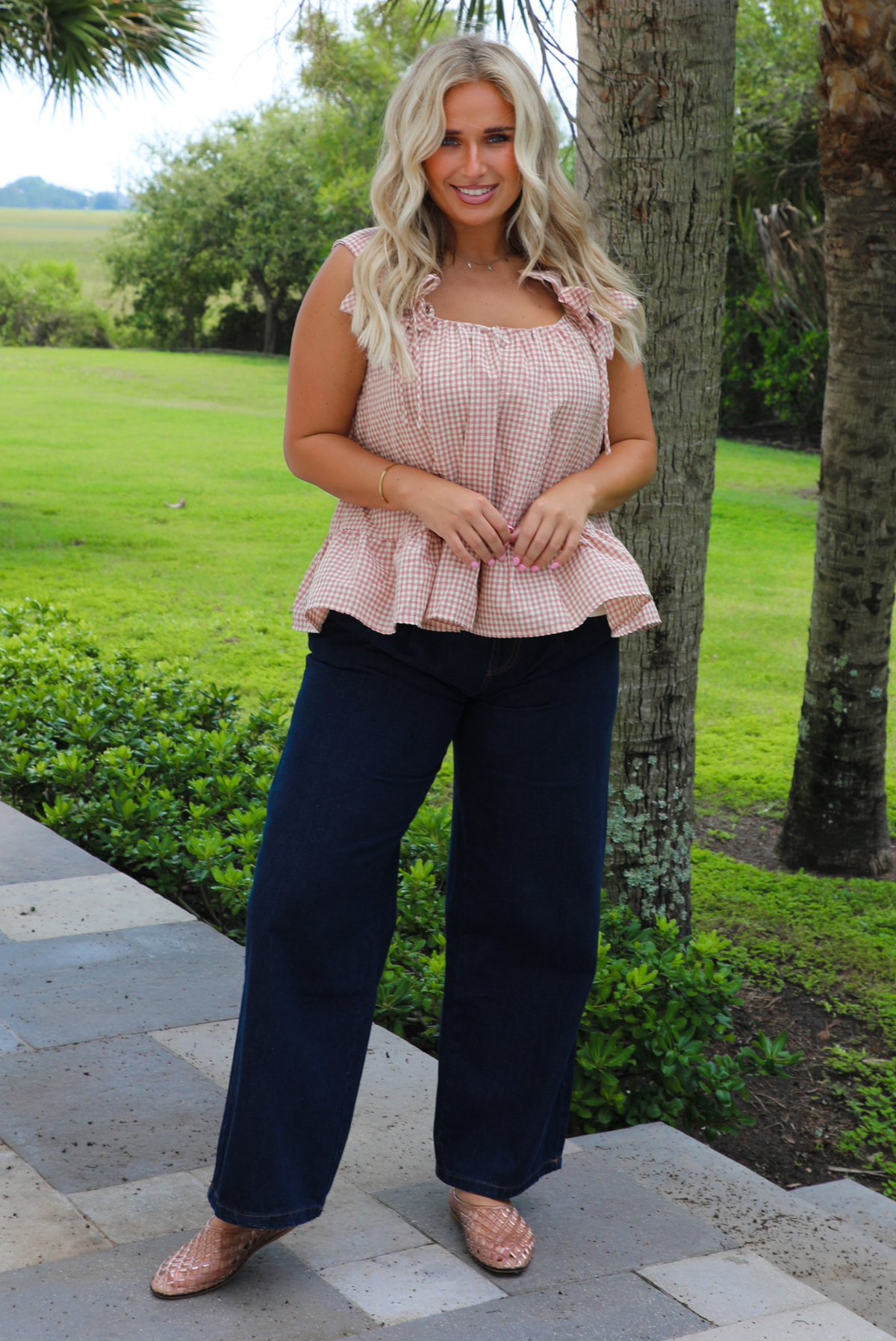 A woman wearing a light maroon and cream gingham flowy top with dark denim jeans, standing on a concrete sidewalk outdoors with palm trees and greenery in the background. 