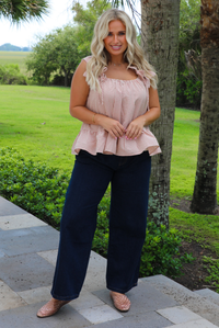 A woman wearing a light maroon and cream gingham flowy top with dark denim jeans, standing on a concrete sidewalk outdoors with palm trees and greenery in the background. 