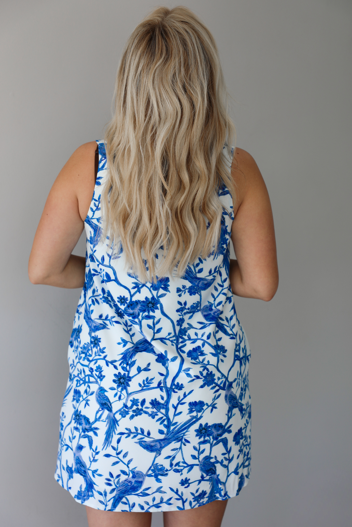 Woman wearing a white and blue print dress with a tree branch and bird pattern throughout indoors against a white wall. The photo shows a closeup of the back. 