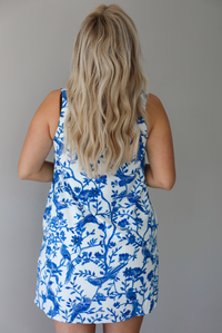 Woman wearing a white and blue print dress with a tree branch and bird pattern throughout indoors against a white wall. The photo shows a closeup of the back. 