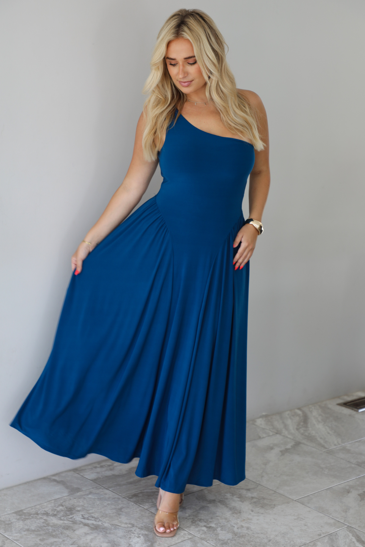 Woman wearing a blue one strap maxi dress indoors against a white wall with white marble flooring. 