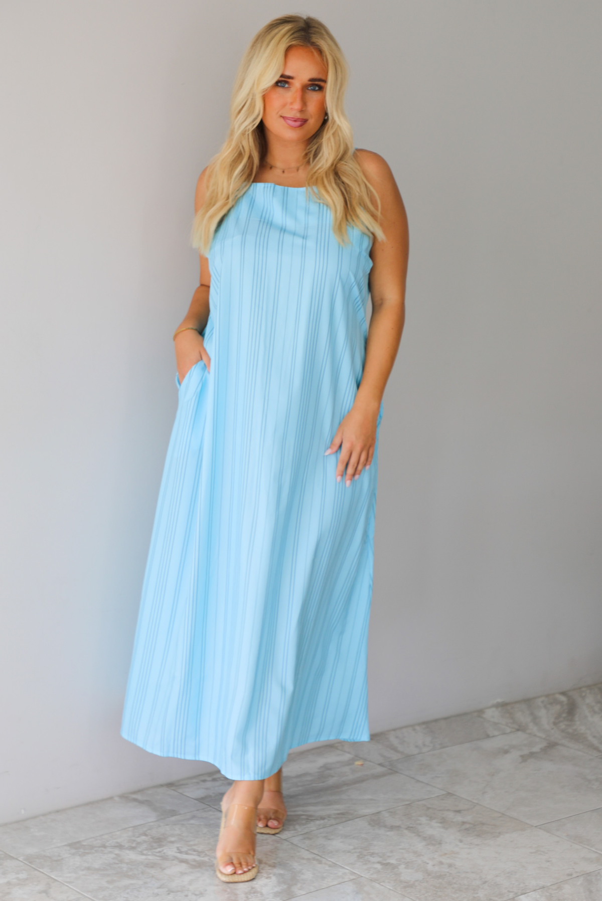 A woman modeling a light blue sleeveless maxi dress with vertical grey pinstripes and side pockets, standing indoors against a white wall with white marble flooring. 