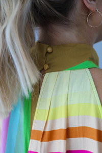 Woman wearing a colorful jumpsuit that has a high neckline, no sleeves, and a flowy cut with flared hem pants. The photo is taken indoors against a white wall and shows a closeup of the button closures in the back at the nape of the neck. 