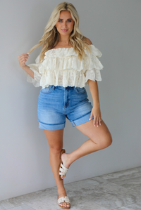 Woman wearing a cream off the shoulder, ruffled eyelet cropped top with medium wash denim shorts indoors against a white wall with white marble flooring. 