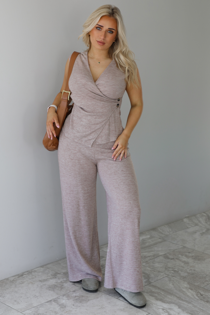 Woman wearing a taupe colored two piece set featuring a vest-appearance top with non-functional button closures on the left side and high waisted pants. It's paired with a tan colored vegan leather shoulder bag. The photo is taken indoors against a white wall with white marble flooring. 