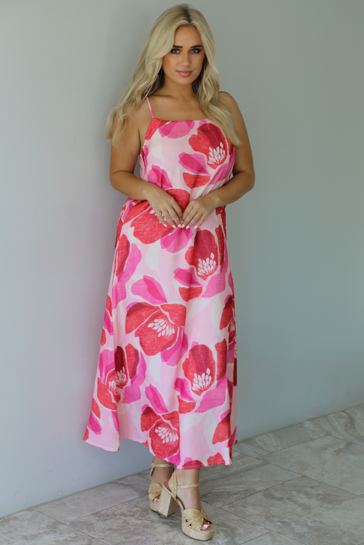 Woman wearing a pink and red floral print maxi dress with thin straps and a flowy fit indoors against a white wall with white marble flooring. 