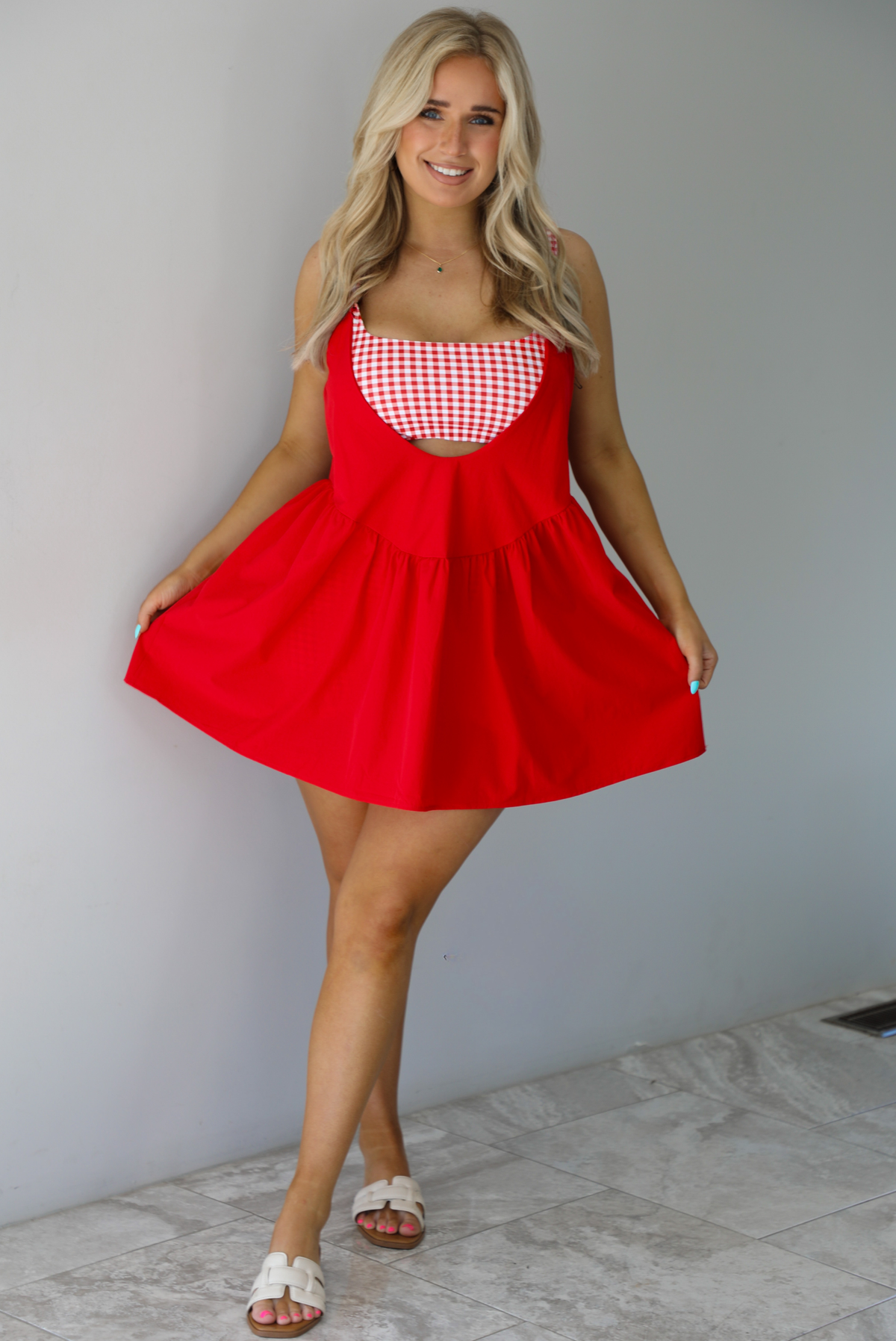 Woman wearing a red dress with a red and white gingham romper underneath. The photo is taken indoors against a white wall with white marble flooring. 