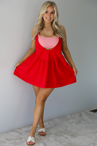 Woman wearing a red dress with a red and white gingham romper underneath. The photo is taken indoors against a white wall with white marble flooring. 