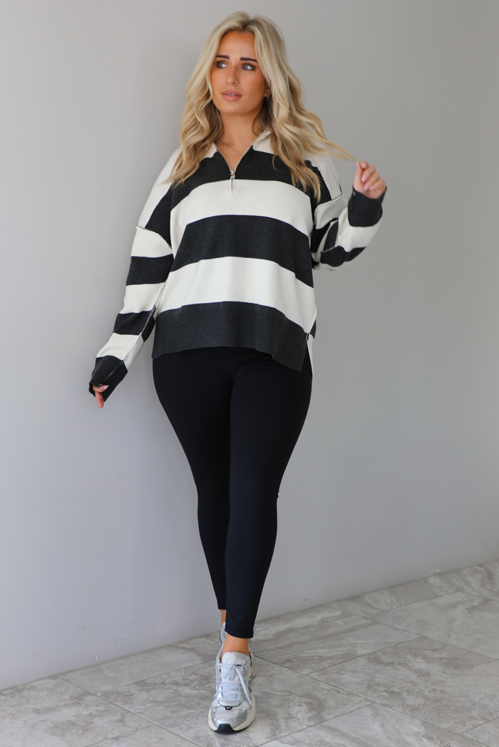 Woman wearing a charcoal and white striped quarter-zip sweater with a collared neckline, raw seam details, and a relaxed fit. It's paired with a fitted black jumpsuit and silver sneakers. The photo is taken indoors against a white wall with white marble flooring.