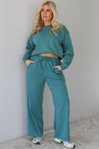 Woman wearing a sage two piece sweat-set. The set features a slightly oversized sweater and high waisted sweat pants that have an elastic and drawstring waistline with pockets. The photo is taken indoors against a white wall with white marble flooring. 