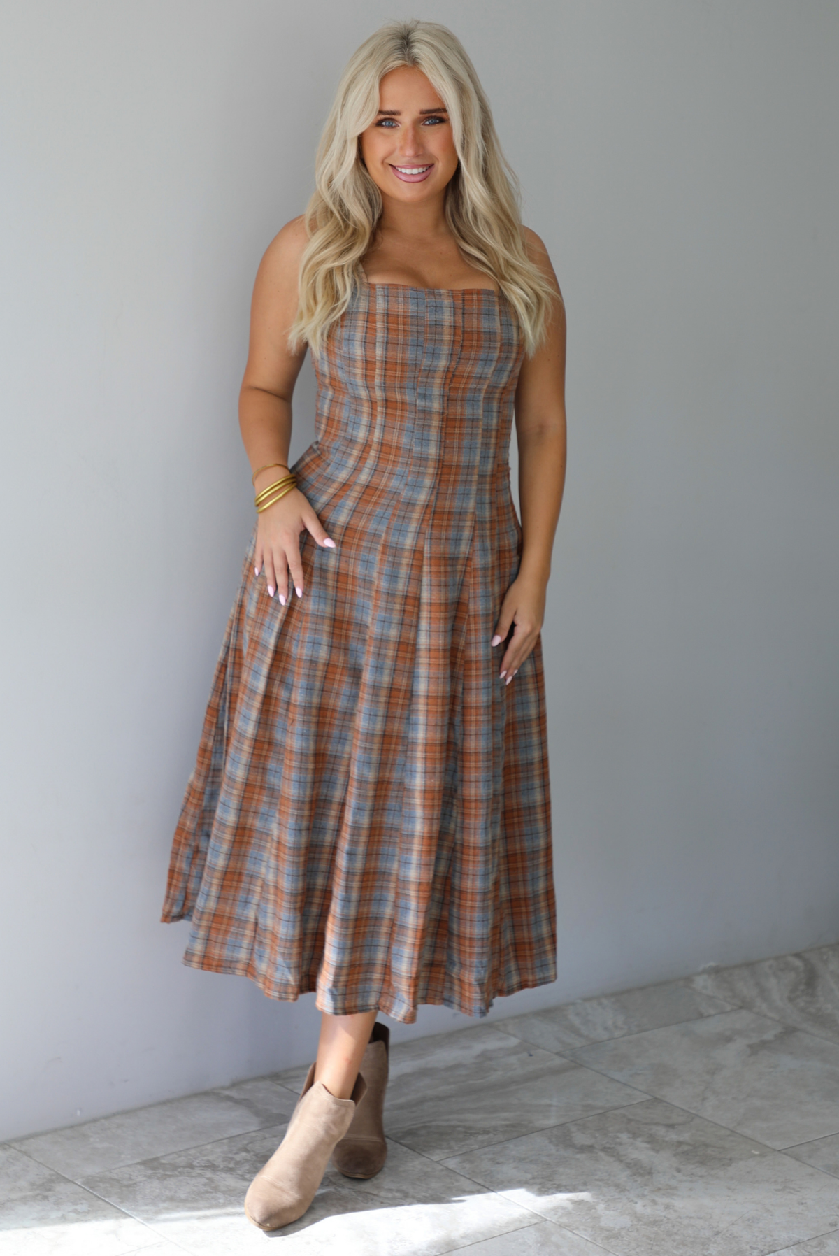 A woman wearing a brown and blue plaid midi dress with thick straps, a square neckline, fitted bodice that starts flaring out at the hips and ends about mid-shin, with a flowy skirt, standing indoors against a white wall with white marble flooring. 