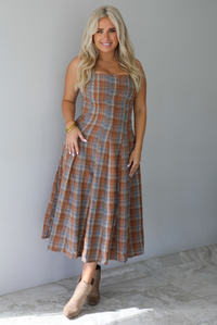 A woman wearing a brown and blue plaid midi dress with thick straps, a square neckline, fitted bodice that starts flaring out at the hips and ends about mid-shin, with a flowy skirt, standing indoors against a white wall with white marble flooring. 