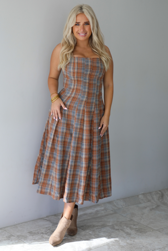 A woman wearing a brown and blue plaid midi dress with thick straps, a square neckline, fitted bodice that starts flaring out at the hips and ends about mid-shin, with a flowy skirt, standing indoors against a white wall with white marble flooring. 
