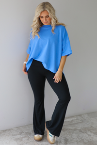 Woman wearing an oversized blue box tee with black leggings that flare slightly at the hem. The photo is taken indoors against a white wall with white marble flooring. 