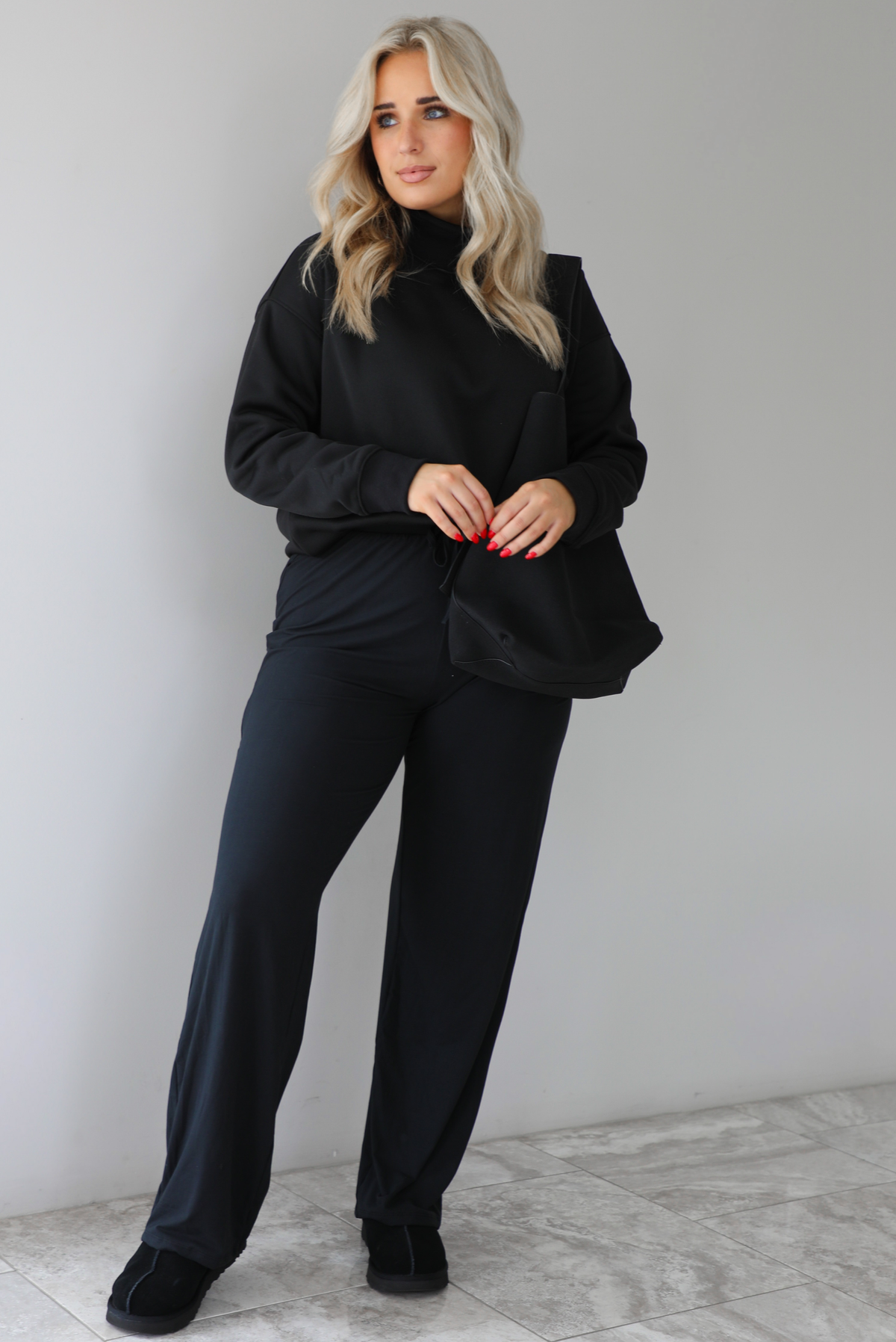 Woman wearing a black high-neck, oversized sweater, high waisted black pants, with a black shoulder bag, and black ugg boot shoes. The photo is taken indoors against a white wall with white marbled flooring. 