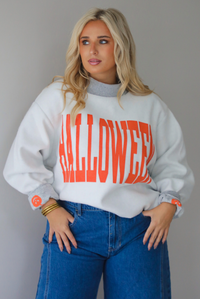 A woman wearing a light grey and dark grey trim oversized sweatshirt that says "HALLOWEEN" in collegiate font on the front, with jack-o-lantern details on the rolled up sleeves, paired with dark wash denim jeans, standing indoors against a white wall.