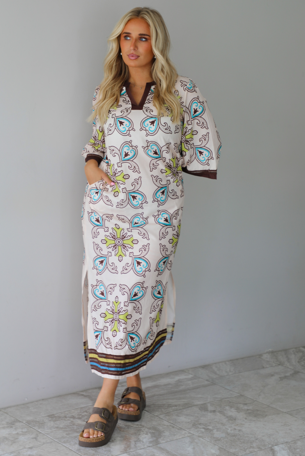 A woman modeling a long kaftan dress with a colorful paisley pattern on a cream background, featuring brown trim and sandals, standing indoors against a white wall with white marble flooring. 