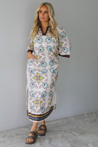 A woman modeling a long kaftan dress with a colorful paisley pattern on a cream background, featuring brown trim and sandals, standing indoors against a white wall with white marble flooring. 