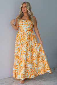 A woman modeling a spaghetti strapped, floor-length maxi dress with a white base and a vibrant marigold orange floral pattern, standing indoors against a white wall with white marble flooring. 