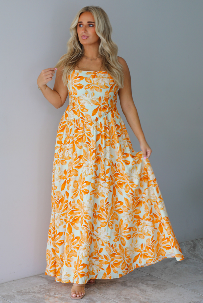 A woman modeling a spaghetti strapped, floor-length maxi dress with a white base and a vibrant marigold orange floral pattern, standing indoors against a white wall with white marble flooring. 