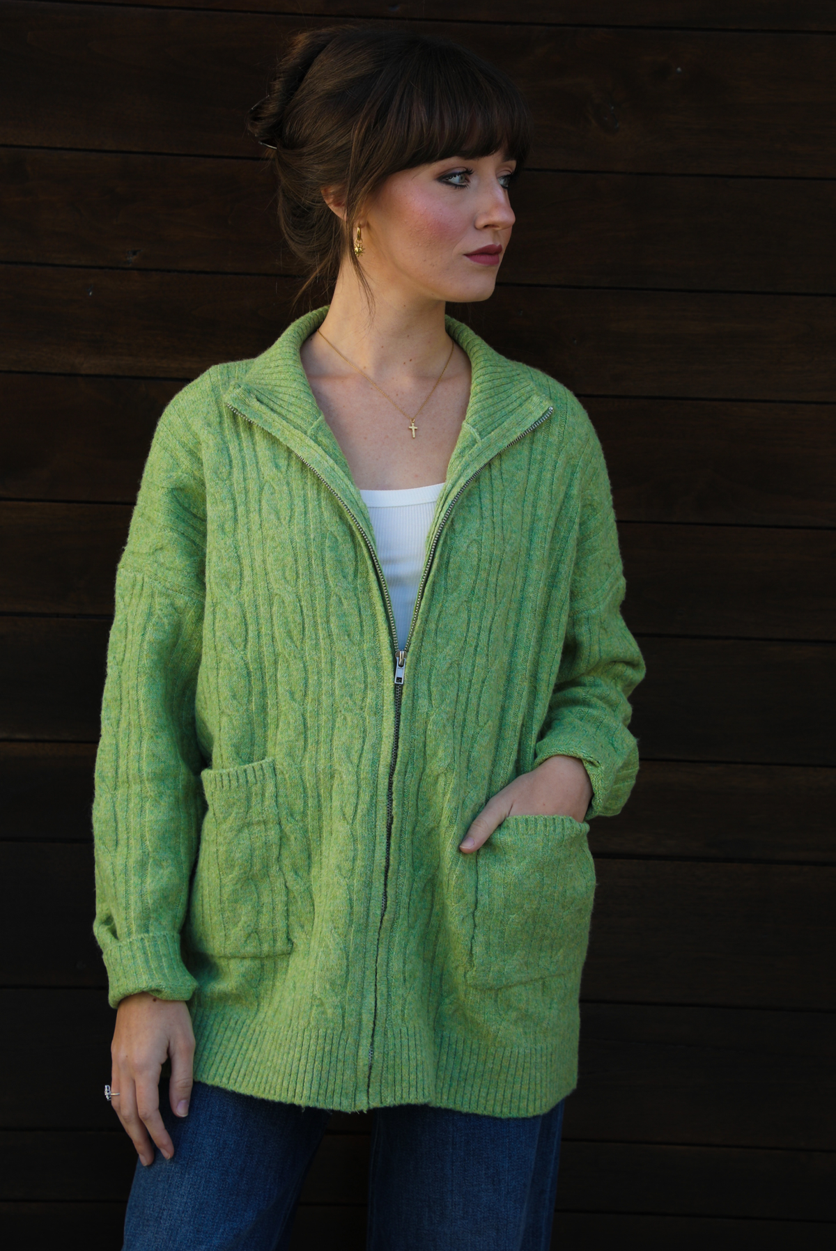 Woman model wearing a green cable knit zip-up cardigan with dark denim jeans outdoors against a dark brown wooden door. The model is wearing a Small. 