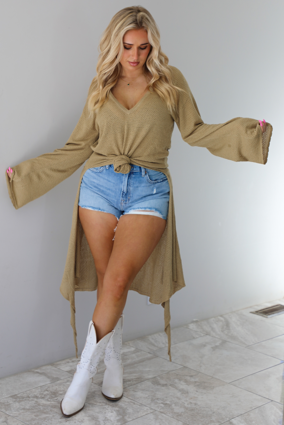 Woman wearing a camel long-sleeve top that's tied in the front with a knot and has a train in the back. The top is paired with denim shorts and white cowboy boots. The photo is taken indoors against a white wall with white marble flooring. 