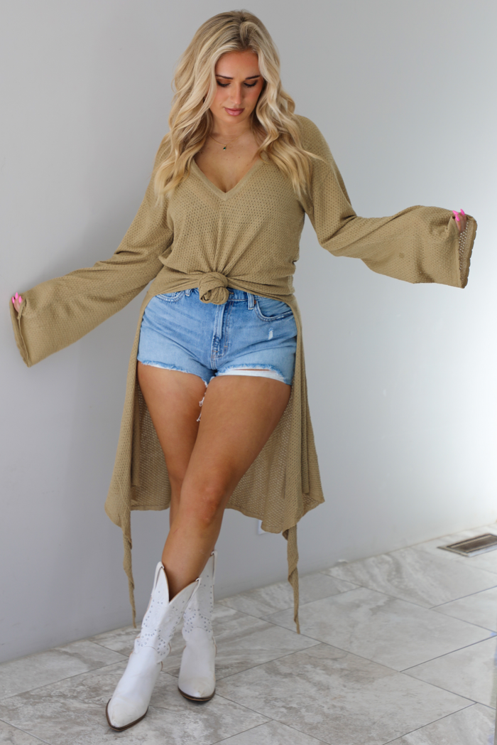 Woman wearing a camel long-sleeve top that's tied in the front with a knot and has a train in the back. The top is paired with denim shorts and white cowboy boots. The photo is taken indoors against a white wall with white marble flooring. 