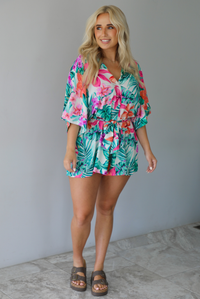 Woman wearing a multi-colored tropical romper with a collared neckline, flowy short sleeves, a cinched with a tie waistline, and flowy shorts indoors against a white wall with white marble flooring. 