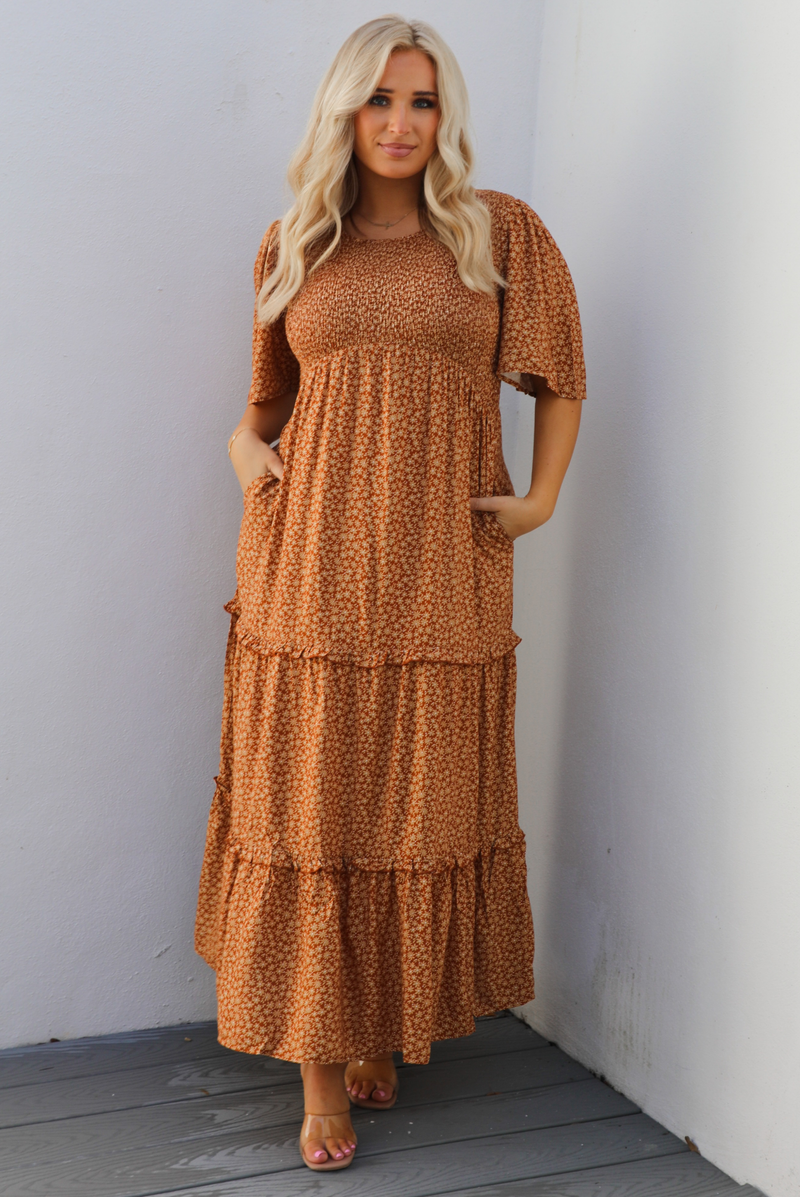 A woman model outdoors wearing a rust dress with a cream floral print pattern throughout. It has flowy short sleeves, a smocked bodice, and a flowy maxi length skirt with functional pockets on either side. 