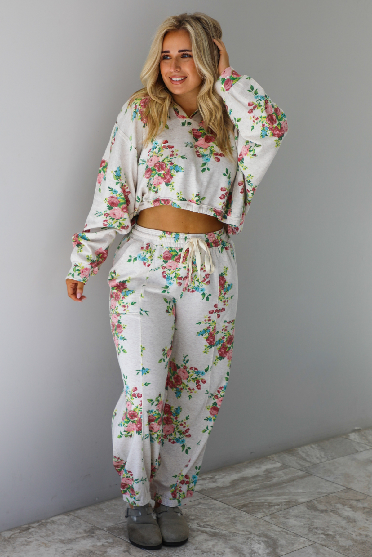 Woman wearing an oatmeal colored and multi-colored floral print two piece set featuring a collared long sleeved crop top and high waisted pants. The photo is taken indoors against a white wall with white marble flooring. 