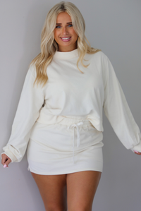 A woman wearing a cream matching two piece set featuring a cropped sweatshirt and mini skirt of the same material, standing indoors against a white wall. The front of the sweater is tucked into the front of the skirt, displaying the drawstring waistline of the skirt. 