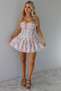 Woman wearing a white and pink floral two piece set featuring a strapless corset style top and an a ruffled skirt. The photo is taken indoors against a white wall with white marble flooring. 