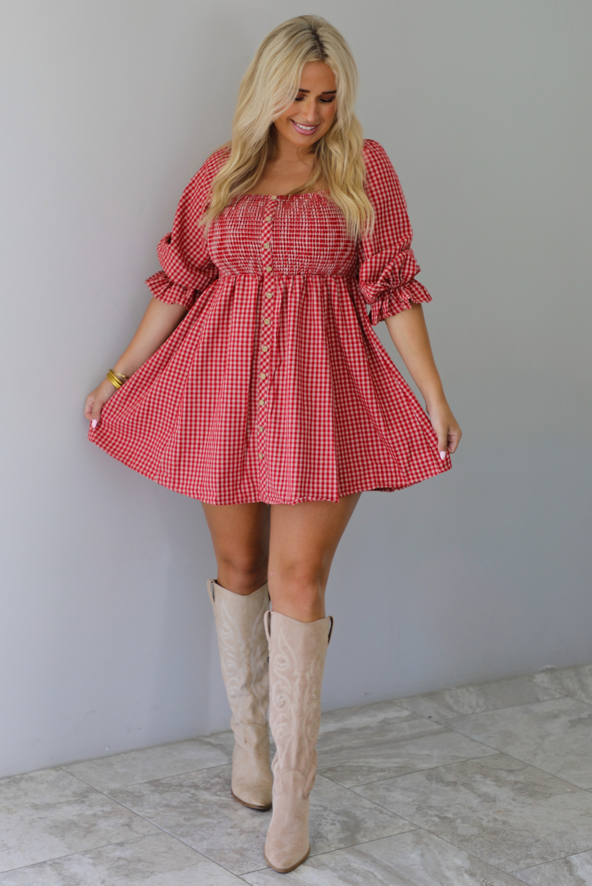 A woman wearing a red and white gingham mini dress with puff, elbow length sleeves and button detailing along the front, paired with beige knee-high boots, standing indoors against a white wall with white marble flooring. 