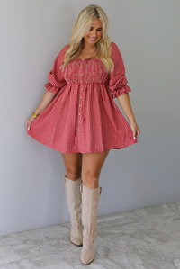 A woman wearing a red and white gingham mini dress with puff, elbow length sleeves and button detailing along the front, paired with beige knee-high boots, standing indoors against a white wall with white marble flooring. 