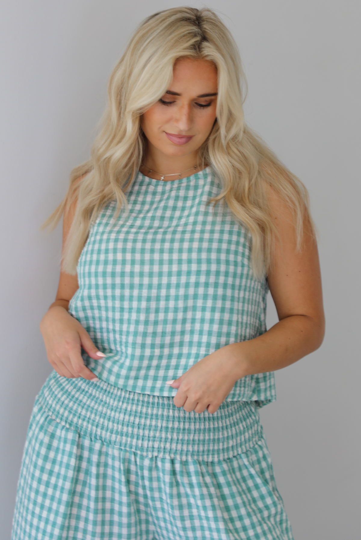 Woman wearing a mint and white gingham set with an oversized top and flared hem pants indoors against a white wall. The photo shows a closeup of the top, which has a high neckline and slightly cropped cut. 