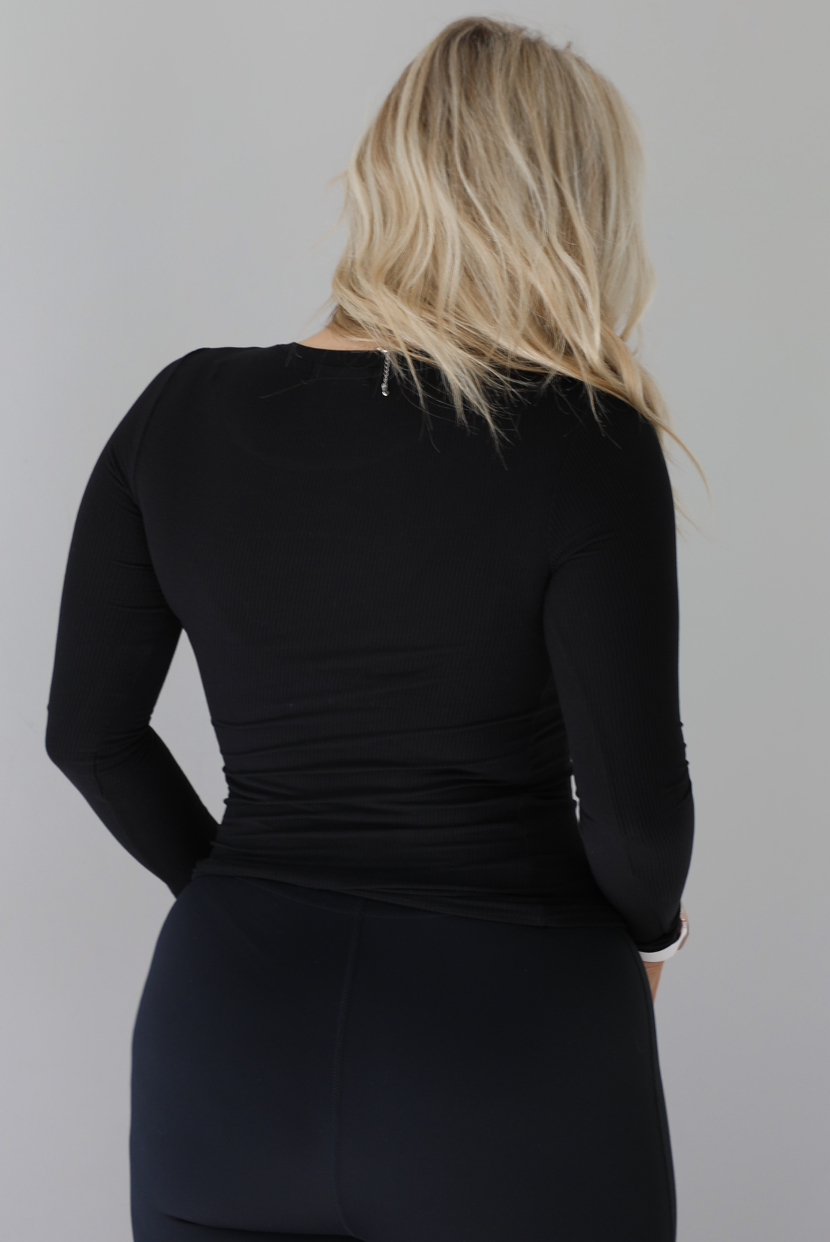 Woman wearing a black, high neckline, long sleeved fitted top that has a ribbed design to the material. It's paired with black leggings. The photo shows the back of the top. The photo is taken against a white wall. 