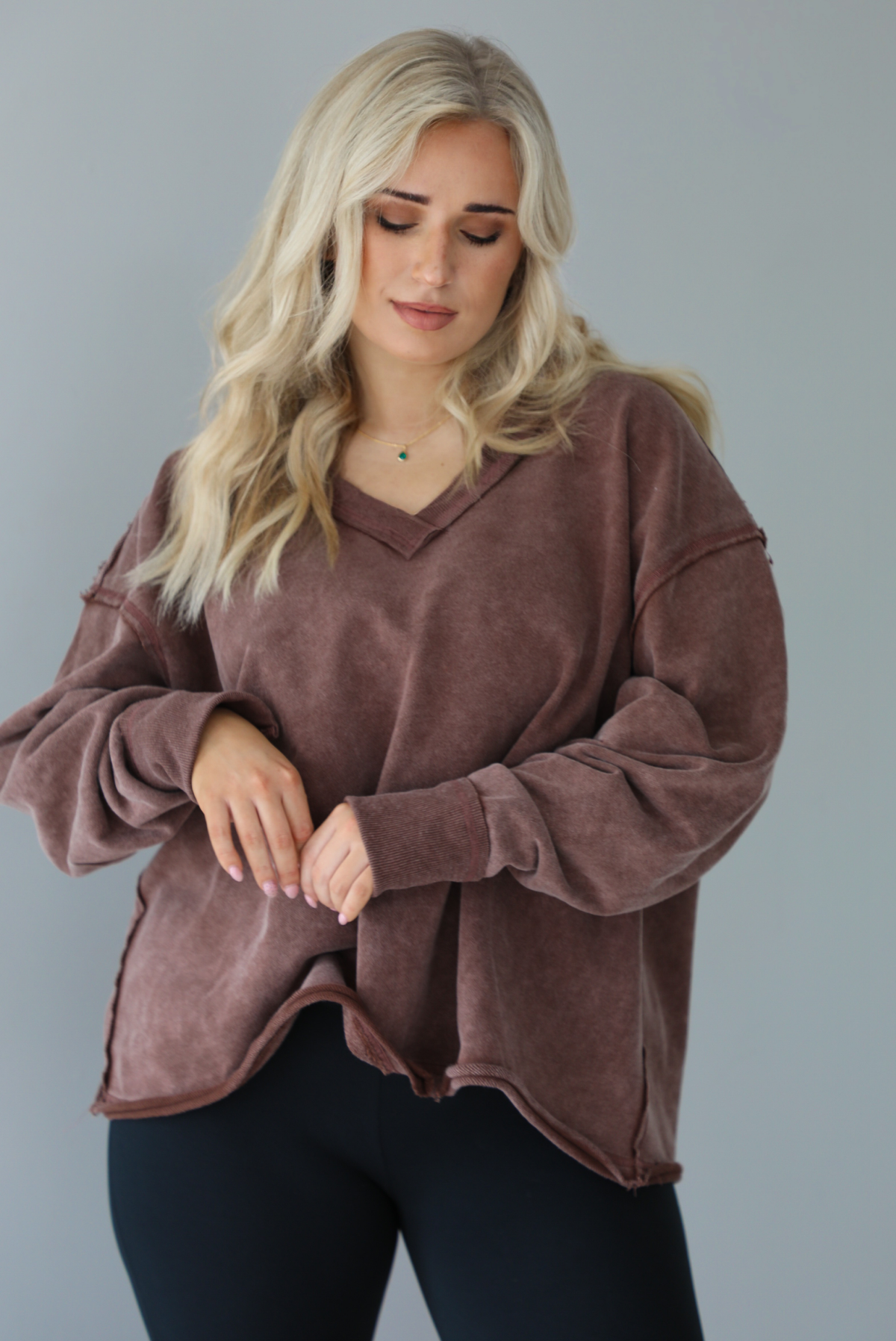 Woman wearing a mauve oversized v-neck sweater with raw hem details with high waisted black flared leggings. The photo shows a closeup of the sweater and the details such as the raw hem and pleated details on the sleeves a the elbow. The photo is taken in front of a white wall.