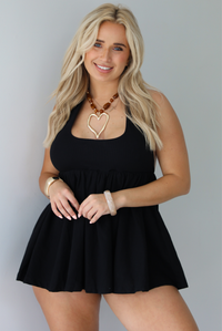 Woman wearing a black romper that looks like a dress, with a halter tie neckline, baby-doll cut, and flowy skirt indoors against a white wall.