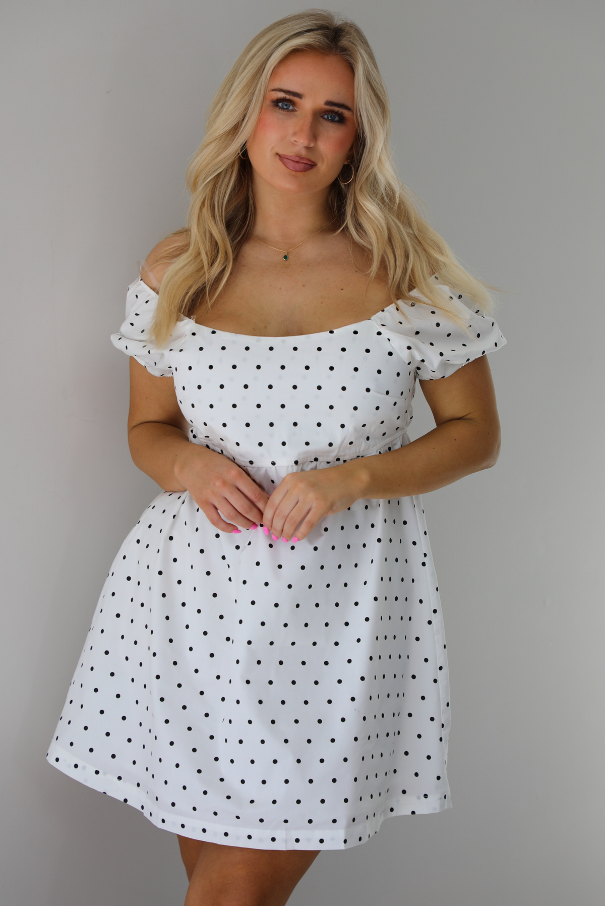Woman wearing a white dress with a black polka dot print throughout. The photo is taken indoors against a white wall.