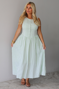 A woman wearing a light green and white gingham maxi dress with a drop waist bodice and side pockets, standing indoors against a white wall with white marble flooring. 