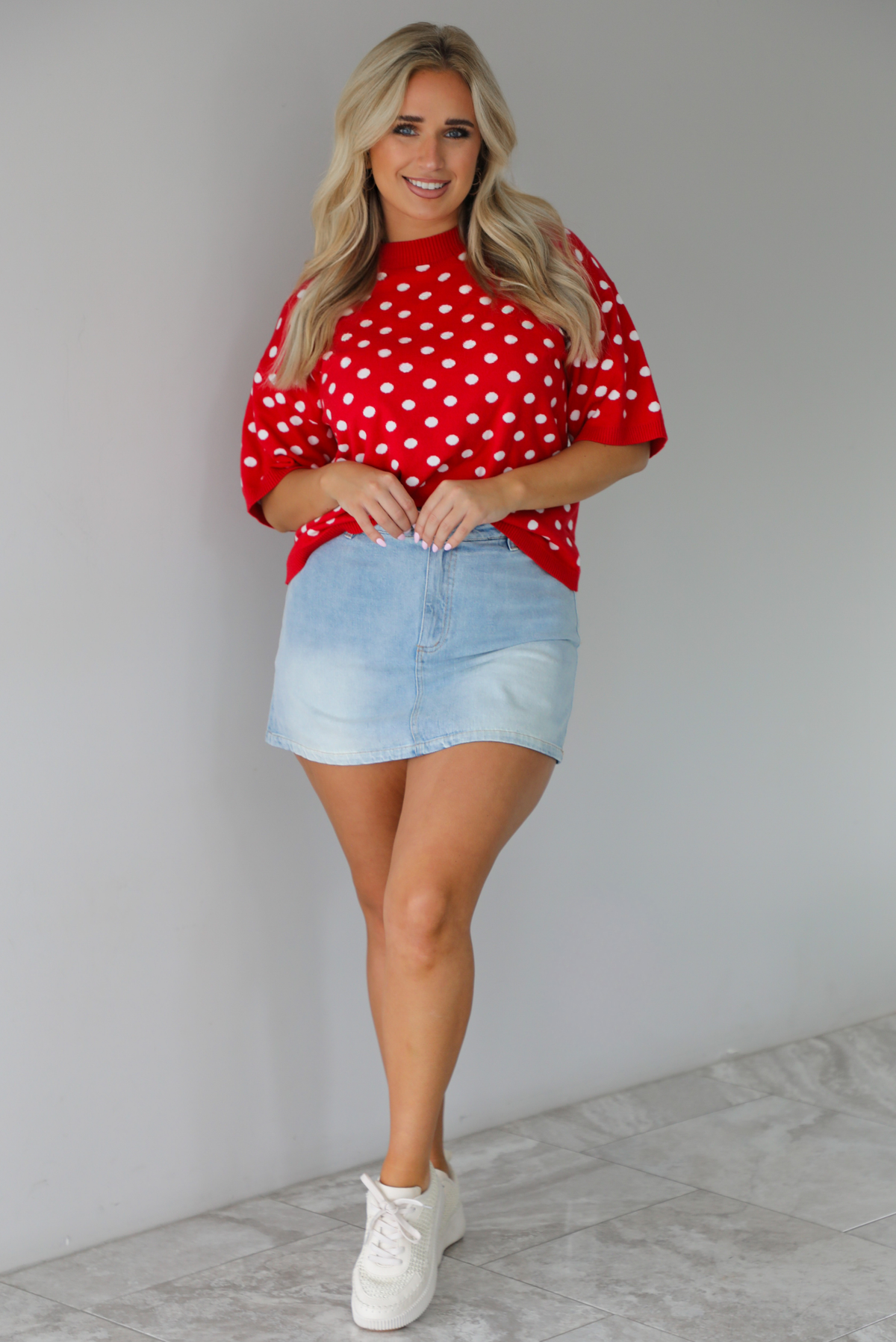 Woman wearing a red knit top with a white polka dot print pattern throughout with a high waisted, light-wash denim skort. The photo is taken indoors against a white wall with white marble flooring. 
